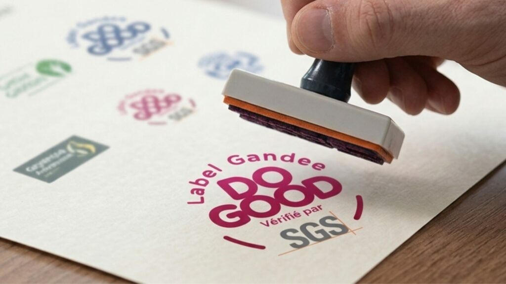 Gros plan sur une main utilisant un tampon encreur pour apposer officiellement le logo "Label Gandee Do Good vérifié par SGS" sur un document.